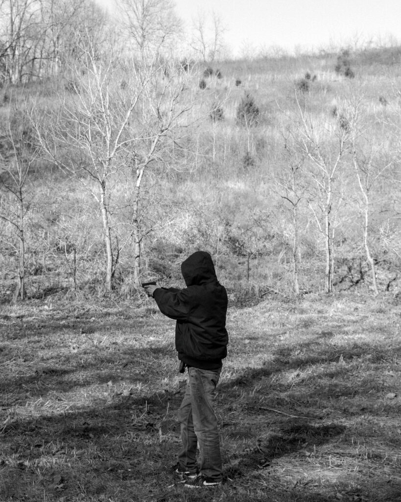 Image: A black and white photograph of a person in a dark hoodie shooting a handgun out in nature. © Rachael Banks. Images courtesy of the artist.