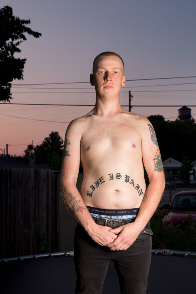 Image: A color photograph of a shirtless man standing facing the viewer. He has tattoos on his arms and a tattoo that says, "LIFE IS PAIN" on his stomach. © Rachael Banks. Images courtesy of the artist.