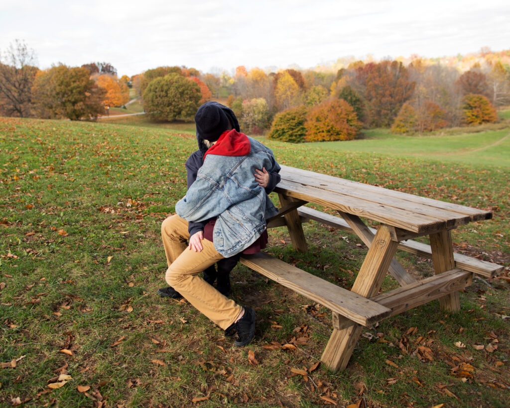 Image: A color photograph of two people sitting next to each other at a wooden picnic table in an intimate embrace. A field of grass and trees are in the background. © Rachael Banks. Images courtesy of the artist.