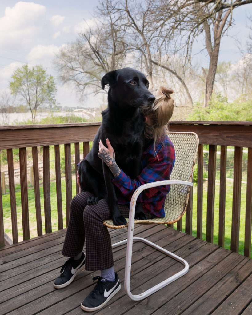 Image: A color photograph of a woman sitting on a chair on a wooden porch with a large black dog in her lap. © Rachael Banks. Images courtesy of the artist.