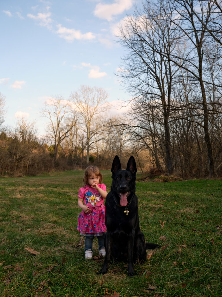 Image: A color photograph of a little girl in a pink dress standing next to a large black dog. The girl has her hand up to her mouth and the dog appears to be smiling. © Rachael Banks. Images courtesy of the artist.