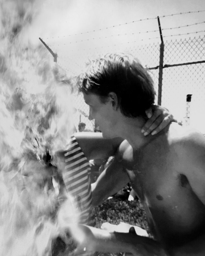 Image: A black and white photograph of a shirtless man embracing a person to our left. The person on the left is obscured. Behind the two people is a chain-linked fence. © Rachael Banks. Images courtesy of the artist.