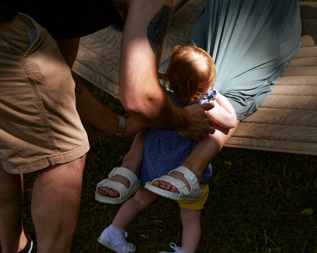 Image: A color photograph of two people with a small child. The viewer's perspective can see the limbs of the two adults and the backside of the small child. © Rachael Banks. Images courtesy of the artist.