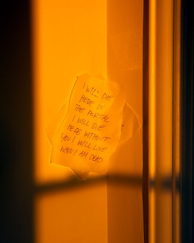 Image: A color photograph of golden light coming in through a window illuminating a section of wall. There is a note on the wall that says, "I will die here in the portal I will die here without you I will live when I am dead." © Rachael Banks. Images courtesy of the artist.