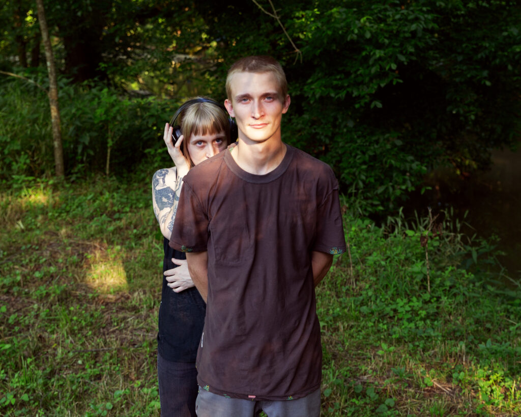 Image: A color photograph of a man in a black shirt facing the camera. A person stands slightly behind him wearing headphones. © Rachael Banks. Images courtesy of the artist.