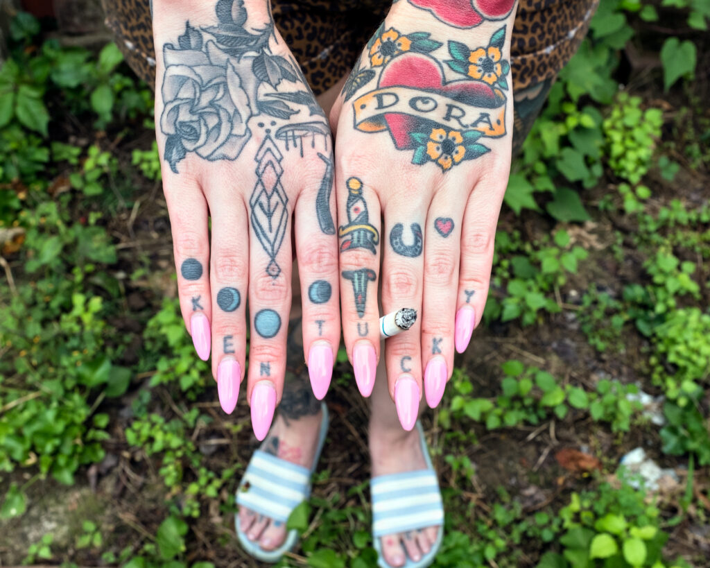 Image: A color photograph of tattooed hands with long pink nails. The hands are extended forward and holding a cigarette. © Rachael Banks. Images courtesy of the artist.