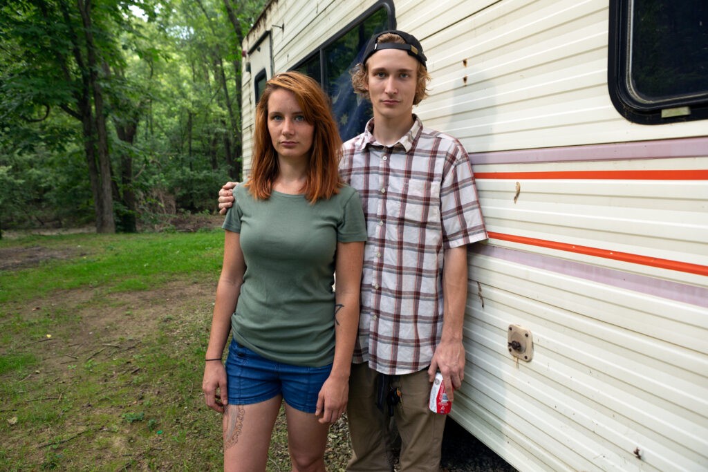 Image: A color photograph of a man and woman standing in front of an RV. The woman has red hair and wears a green shirt and shorts. The Man has blond hair and wears a backwards cap and a plaid shirt. © Rachael Banks. Images courtesy of the artist.