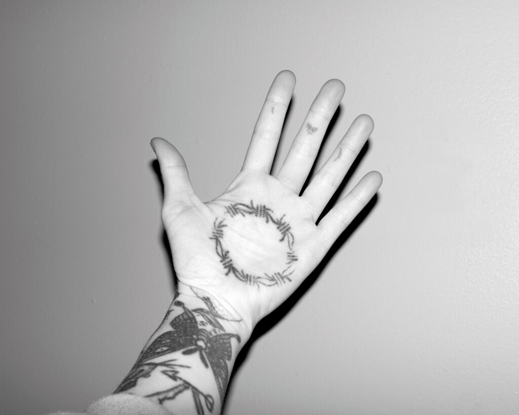 Image: A black and white photograph of a person's hand extended with their palm out. The persona's arm has several tattoos, and the palm of the hand has a tattoo of a ring of barbed wire. © Rachael Banks. Images courtesy of the artist.