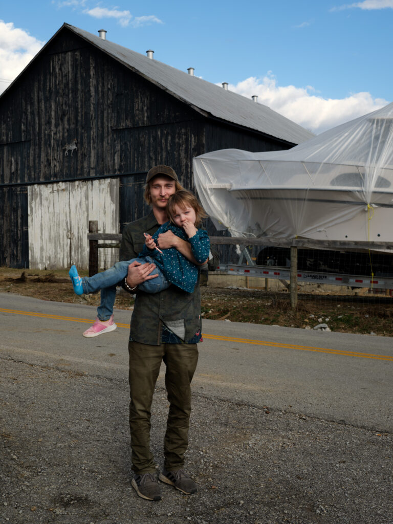 Image: A color photograph of a man standing facing the camera while holding a young child. He is standing on a road in front of a wooden barn next to a covered boat. © Rachael Banks. Images courtesy of the artist.