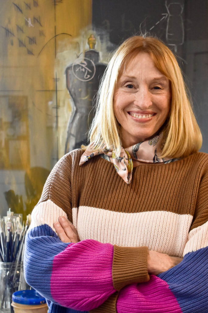 Image: A photograph portrait of the artist, Honora Jacob, in her studio. She is wearing a multi-colored sweater and a pink silk scarf. She smiles and looks directly at the viewer. Photo by Kevin Nance. 
