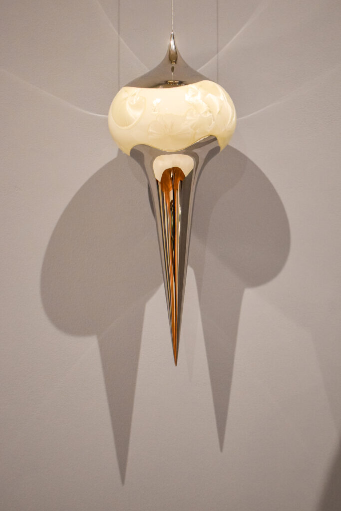 Image: Li Hongwei, Xuan #41 (detail), porcelain and stainless steel, 23.5" x 8.75" x 8.75”. A silver and cream colored sculpture hangs in front of a grey wall. The sculpture is mostly round on top, but forms a point at the very top. The shape of the sculptures comes down into a point at the bottom. Photo by Kevin Nance.
