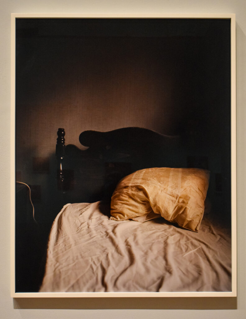 Image: Todd Hido, untitled image from House Hunting (2019), pigment C-print. The piece depicts a bed with off-white sheets and a yellow pillow in a shadowy bedroom. Photo by Kevin Nance.