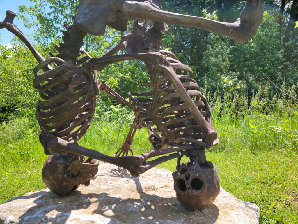Image: Upsidedown HoDown by Araan Schmidt. A photo of a bronze sculpture of two skeletons intertwined and inverted on a rock.