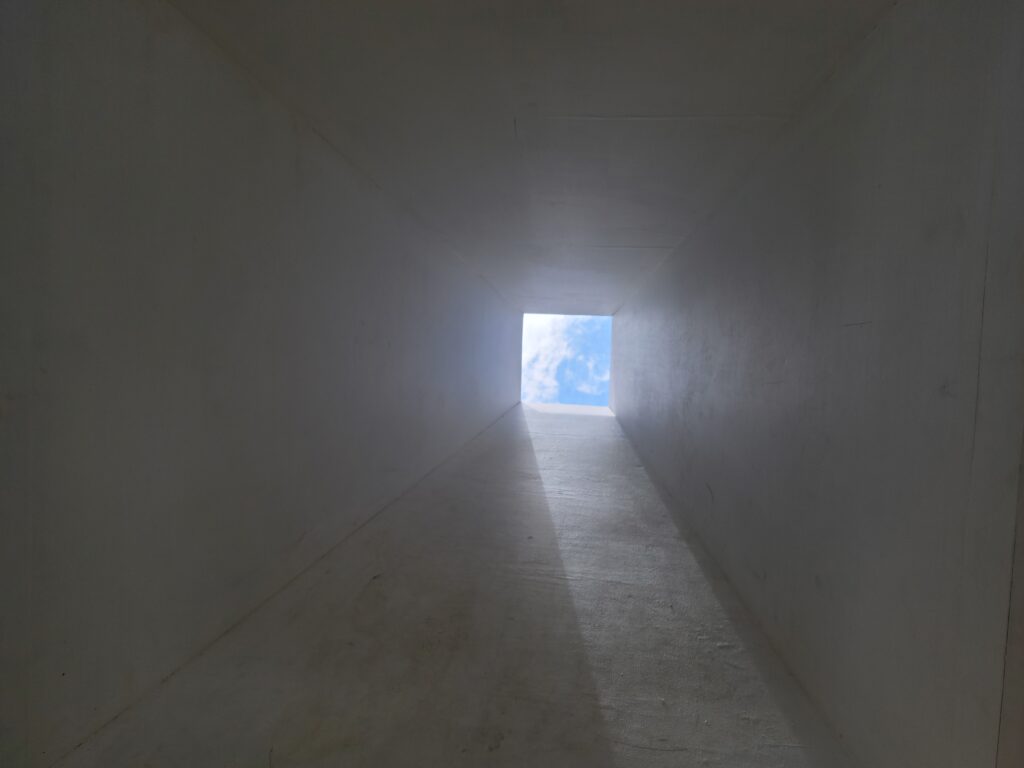 Image: A photo of the inside of the wooden structure after crawling into the square opening. The inside shows a long square hole with another opening at the end, revealing a square patch of blue sky.
