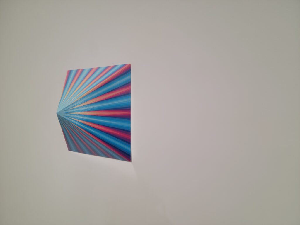 Image:  Gibbs Rounsavall, Interface #4 (Peak), 2022, enamel on wood panel. A three-dimension, pink and blue piece hangs on a wall. The piece is shaped like a pyramid and comes out into a point. Photo by the author.
