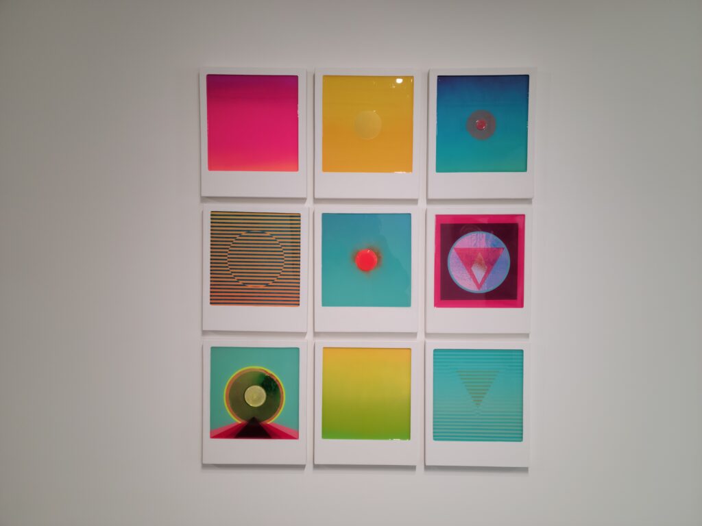 Image: Letitia Quesenberry, As of Yet, 2020-2022, polished plaster, panel, paint, resin. Nine colorful square pieces showing various geometric shapes are on display in a grid on a white wall. Photo by the author.