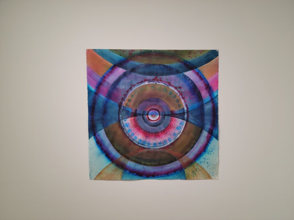 Image: Skylar Smith, Water, watercolor, ink, acrylic, gouache, colored pencil on paper, 2022. An abstract piece of various circle shapes in shades of blues, greens, pinks, and oranges hangs on a white wall. Photo by the author.