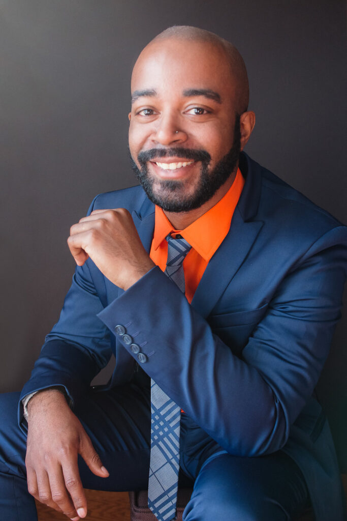 Image: A photo of Joe Tolbert, Jr., Knoxville, who is the program director of the Waymakers Collective facilitation team. His background is in cultural work and he runs Arts the the Intersections. He is smiling at the viewer while wearing a blue suit and an orange shirt. Photo provided by the Waymakers Collective.