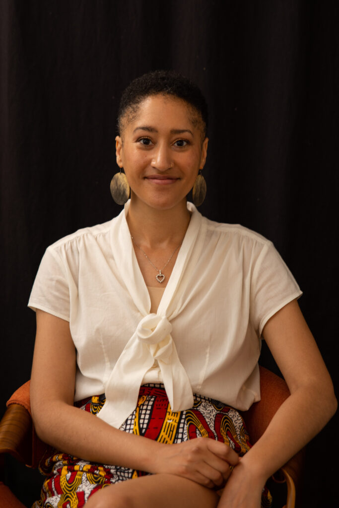 Image: Dr. fari nzinga, Curator of Academic Engagement and Special Projects at the Speed Art Museum in Louisville, Kentucky. nzinga sit in a chair while smiling at the camera. She is wearing a white blouse and a red and yellow skirt. Photo credit: Aja Joi.