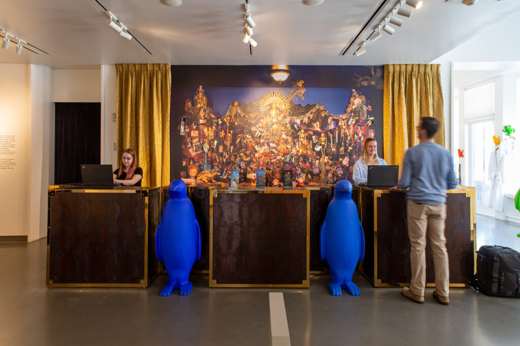 Image: Robert Morgan, digital reproduction of Avalanche, 1996 and ongoing Mixed Media Alterpiece, at 21c Lexington. The photo shows the front desk area at 21c, where three people stand amongst two large blue penguin statues. On the wall behind them is a nearly life-size, photographic replica of a wall in Morgan’s home with an assemblage-like altar. Image courtesy of 21c Lexington.