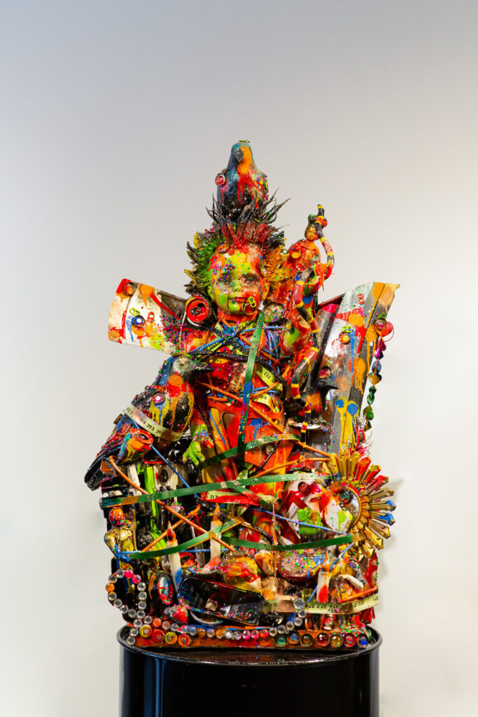 Image: Robert Morgan, Feeding Crows, 2021. Mixed Media. A colorful, assemblage sculpture with a doll's head on top sits on a black pedestal. Image courtesy of 21c Lexington.