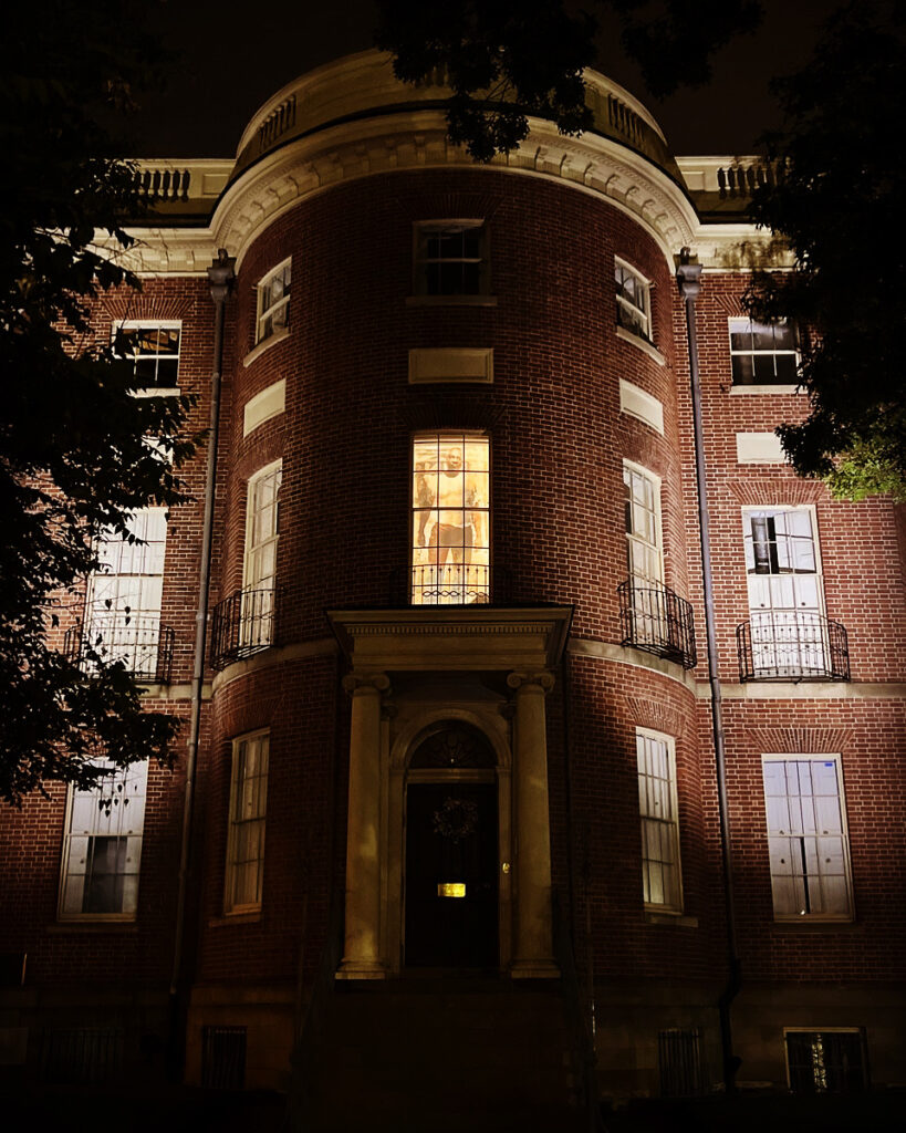 Image: Installation for the I Was Here project, courtesy of Marjorie Guyon. A large photo of a man can be seen from the outside of a brick building through a window.