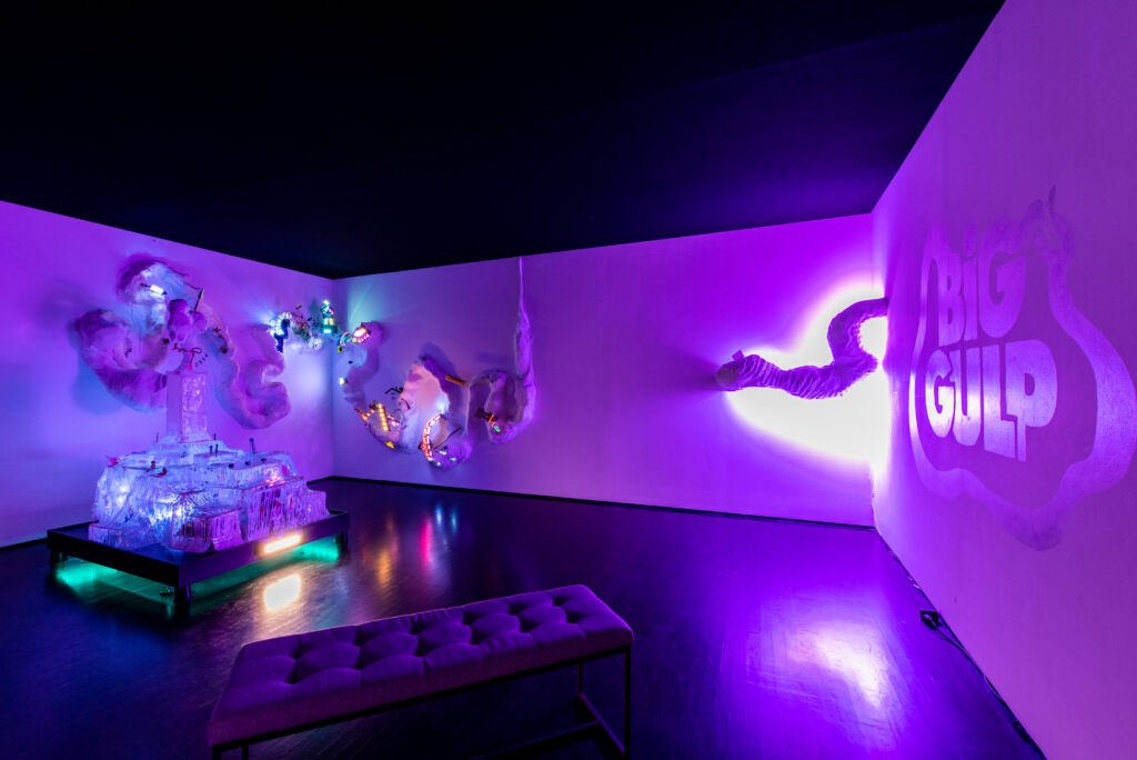 Image: Detail of Hannah Smith, Big Gulp (2022). Thermoformed plastic, animated LED circuit, programmed hologram, papier-mâché, laser-cut chipboard, UV printed plexiglass, fiberoptics, automatic bubble blower. Dimensions vary. The installation is made up of various shapes, but are mostly hues of violets and blues due to the lighting in the installation. On the right, the wall says the words "BIG GULP". Image courtesy of the artist.