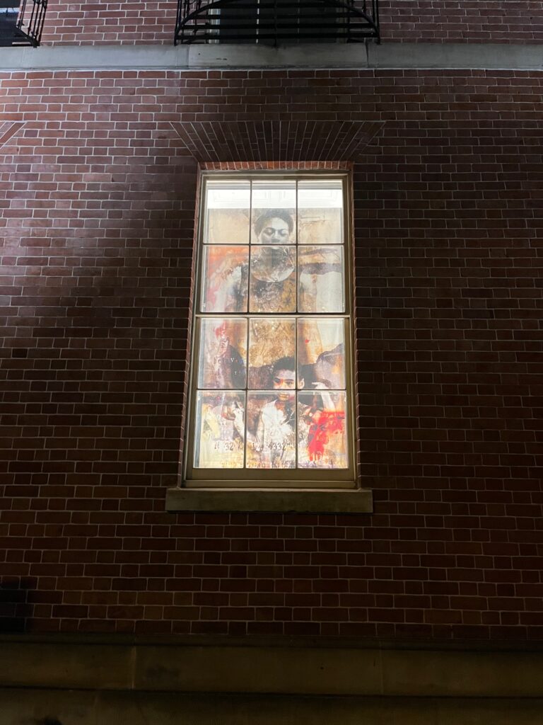 Image: Installation for the I Was Here project, courtesy of Marjorie Guyon. A large photo of a very young boy and his mother can be seen from the outside of a brick building through a window.