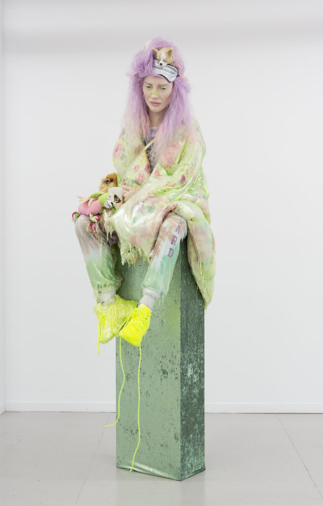 Image: Cajsa vonZeipel, Pack Nap, 2019. Mixed media and silicone. Courtesy of Laura Lee Brown and Steve Wilson, 21c Museum Hotels. A sculpture of a person with purple hair wearing comfy clothing sits on a green pedestal. She wears a blanket wrapped around her body.