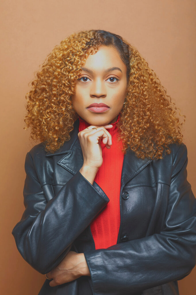 Image: A photograph of Alivia Blade. She is wearing a leather coat over a red top. On hand rests thoughtfully on her chin while she looks forward towards the camera.