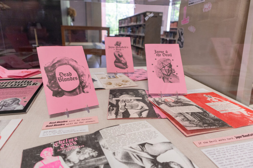 Image: Claire Thompson, Jayne Mansfield's Head, Jayne & the Devil, and Dead Blondes, 2022, 5" x 7", three Risograph-printed zines, from The Jayne Mansfield Cycle series. Three pink zines are propped upright on display, while others lay open on a surface. Photo by author Josh Porter. 