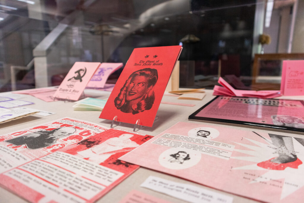 Image: Installation view of Beautifully Grotesk at the Lucille C. Little Fine Arts Library. The photo shows a selection zine laying open on display with several others propped upright. The zines are mostly red, pink, and white. Photo by author Josh Porter. 