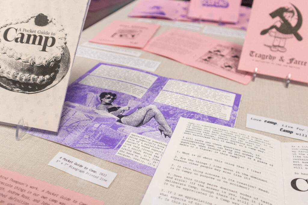 Image: Claire Thompson, A Pocket Guide to Camp, 2022, 5" x 7", Risograph-printed zine. On display are several open zines with two zines propped up. The zines are mostly purple, pink, and white. Photo by author Josh Porter. 
