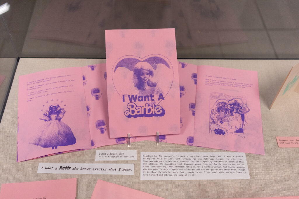 Image: Claire Thompson, I Want A Barbie, 2022, 5.5x8.5, risograph printed zine. A display of pink zines lay open on a surface with purple images of Barbie inside. The cover of the zine shows Barbie and says, "I Want A Barbie". Photo by author Josh Porter.