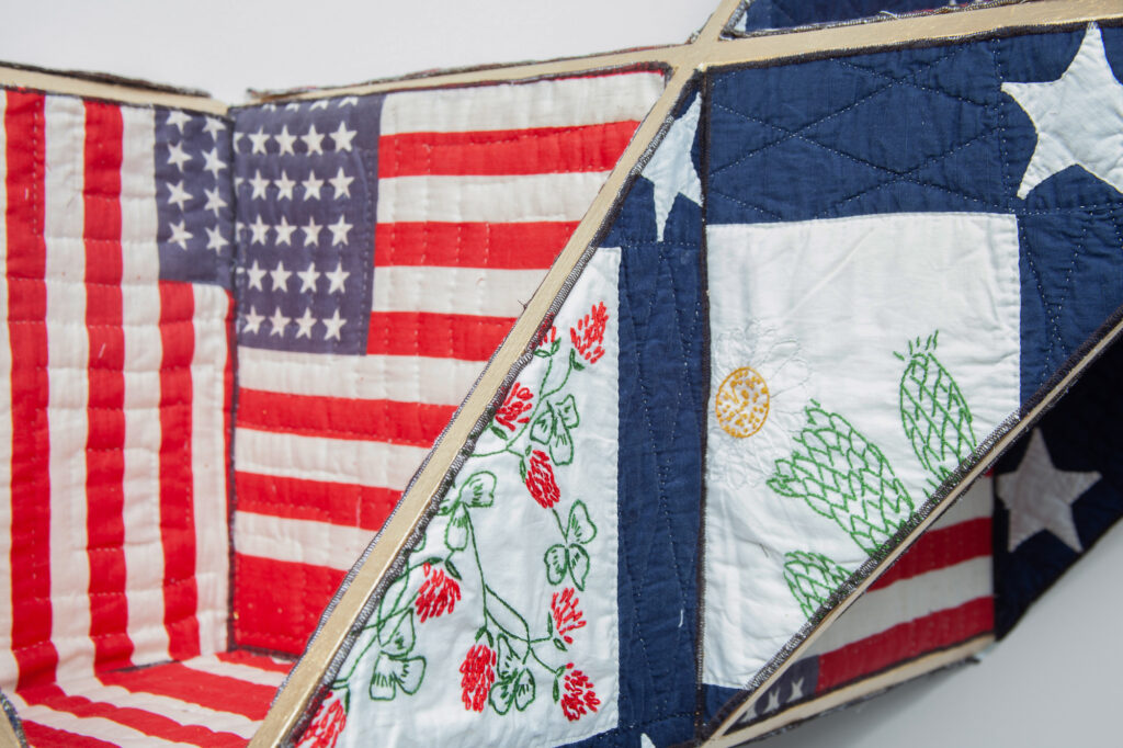 Image: A detail view of Reconstruction, 2019 by Sanford Biggers. Antique quilt, birch plywood, gold leaf. 38 x 72 x 19 in. Marty Nesbitt (Collector) Chicago, IL. Original quilt used: ssbqmat092. Courtesy of the artist and Monique Meloche Gallery, Chicago. Photo: RCH Photography. A quilt with American flag patterns reconstructed on wood so that it takes on a three-dimensional shape.