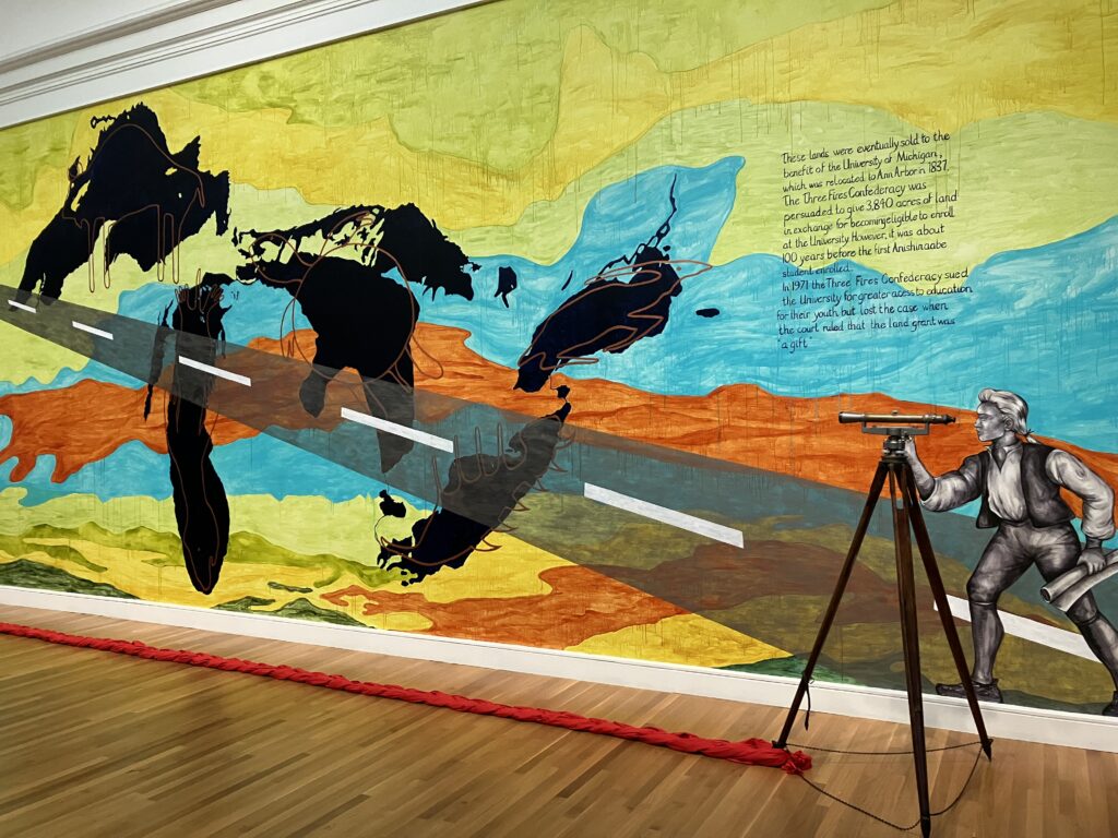 Image: Bonnie Devine, The Gift, 2022. Acrylic paint, graphite, ink, beaded felt, surveyor’s transit, and red cotton cloth. A large mural with text and a map of the Great Lakes can be seen on a wall. In front, a tripod with a telescope is set up with a red cloth tied around its base. Photo by Kim Kobersmith.