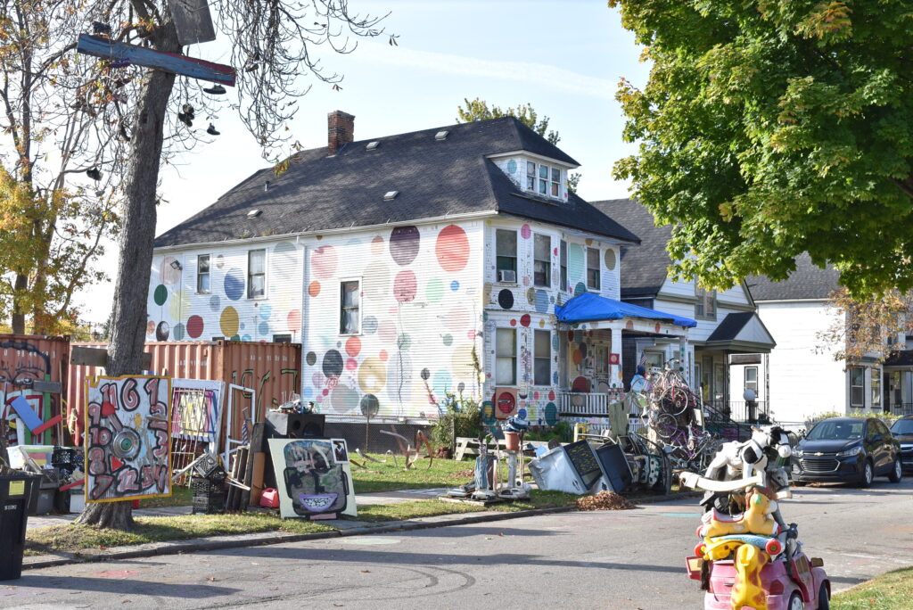 Image: Artist Tyree Guyton’s childhood home is now known as the Dotty Wotty House and is an iconic symbol of the project. An assortment of sculptures surrounds the house: hanging shoes, a tower of bicycles, stacked plastic riding toys, and a series of old doors leaning against shipping containers. Photo courtesy of the author.