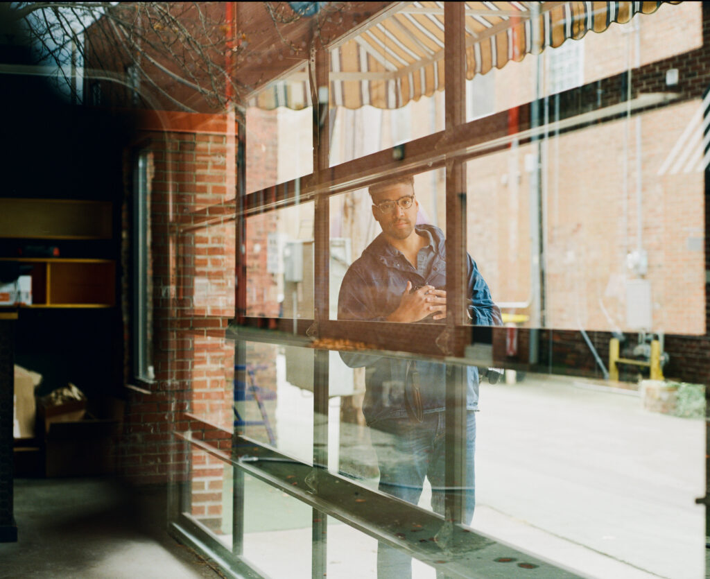 Image: A photo of Jon Cherry standing outside of a transparent garage-like door. He is wearing mostly denim and also wears glasses. He look directly at the viewer. Image courtesy of the artist.