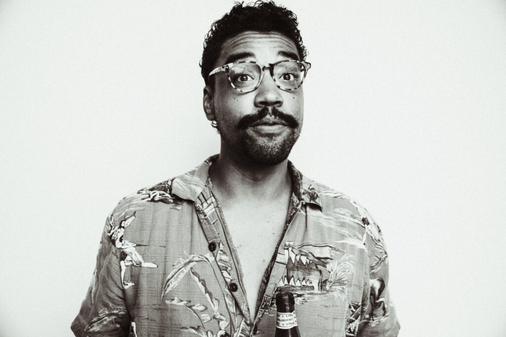 Image: A black-and-white photo of Jon Cherry. He is wearing a button up shirt and glasses while looking directly on the viewer with his eyebrows raised. Image courtesy of the artist. 