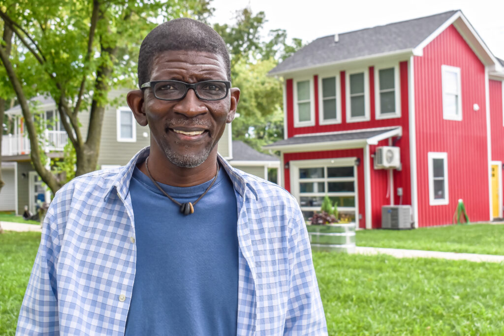 Image: A photo of Mark Lenn Johnson at his home and studio at the Artists' Village. He stands in front of a bright red house while smiling at the camera. Photos by Kevin Nance.
