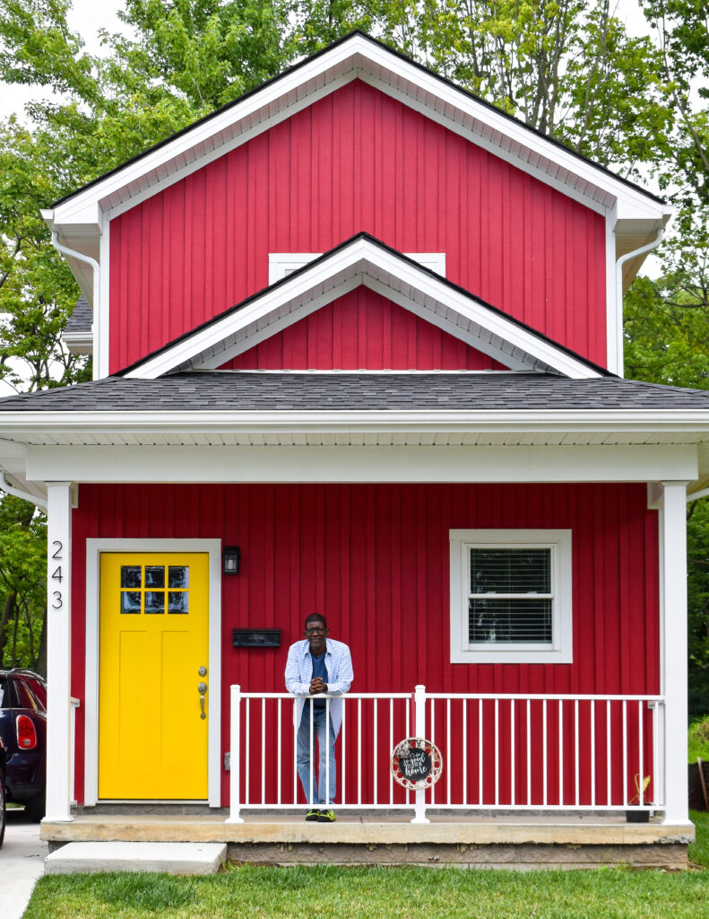 Image: Mark Lenn Johnson at his home and studio at the Artists' Village. He stands on the porch with his hands folded while facing the camera. The house behind him is bright red with a yellow door. Photo by Kevin Nance.