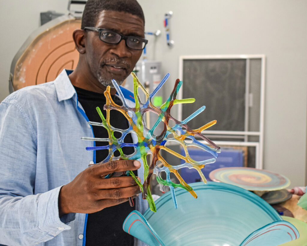 Image: Mark Lenn Johnson, seen here in his home studio holding a colorful glass piece, likes to experiment with color and overlapping patterns to design his glass bowls. Photo by Kevin Nance.