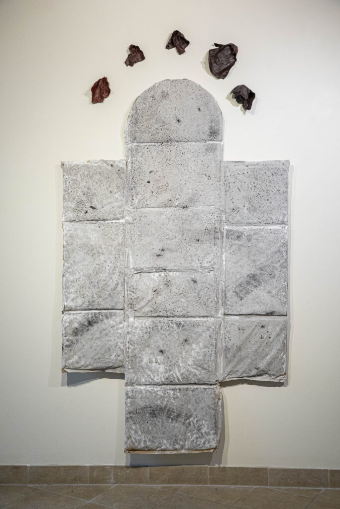 Image: House of Slaves by Lisa Bachman Jones. A sculptural piece that looks like a stone floor hangs on a wall. It is mostly comprised of grey rectangles, with the top part shaped in an arch. Above the arch, five dark organic-shaped pieces are hung in an arch shape. Photo by Annaliese Loughead, courtesy of The Parthenon Gallery.