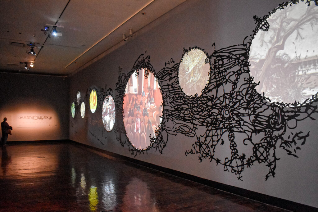 Image: Installation view of Matthew Ritchie's piece titled The Arguments, 2022. Ten-channel multimedia installation with sound database, acrylic, and ink. Projections of images in the shape of circles can be seen displayed on a long wall. The projections are framed by black markings. Photo by Kevin Nance.