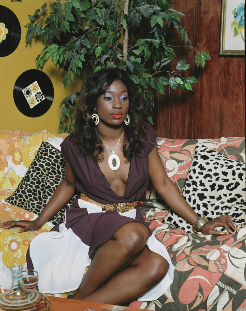 Image: Mickalene Thomas, Portrait of Qusuquzah, 2008. Chromogenic print. A photograph of a woman sitting on a multi-patterned sofa. She places her hands on the sofa on either side of her and looks slightly towards the viewer's right. Courtesy of 21c Museum Hotel, Nashville.